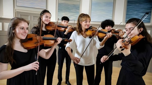 Schüler der St. Mary's Music School spielen gemeinsam Geige in einem Raum mit großen Fenstern und Blick auf ein Gewässer.