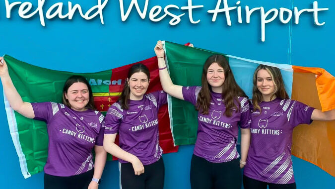 Vier Schülerinnen der St. Mary’s Secondary tragen lila Trikots und halten Fahnen vor dem Ireland West Airport.