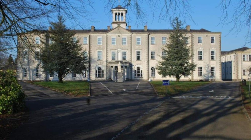 Das mehrstöckige Gebäude mit markantem Turm auf dem Gelände von St. Mel’s College ist von Bäumen und einem gepflasterten Bereich umgeben.