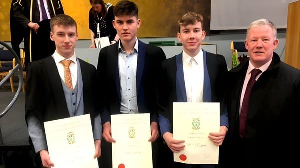 Absolventen von St. Mel’s College im formellen Outfit halten ihre Diplome während der Abschlussfeier mit einem Ehrengast.