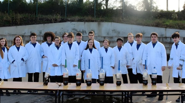 Eine Gruppe Schüler in weißen Laborkitteln steht auf dem Gelände von St. Michael's College vor einer Betonwand mit Bäumen im Hintergrund.