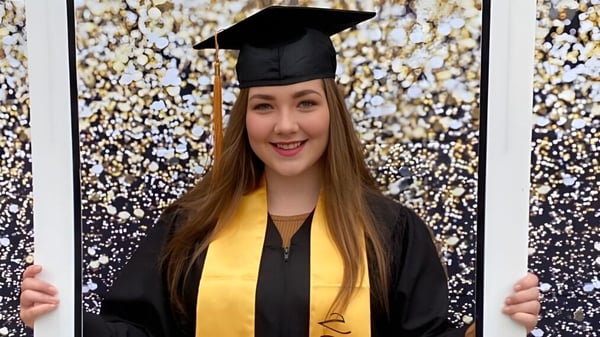 Eine junge Frau in Abschlussrobe steht vor einem glitzernden Hintergrund auf dem Campus von St. Michael's Regional High.