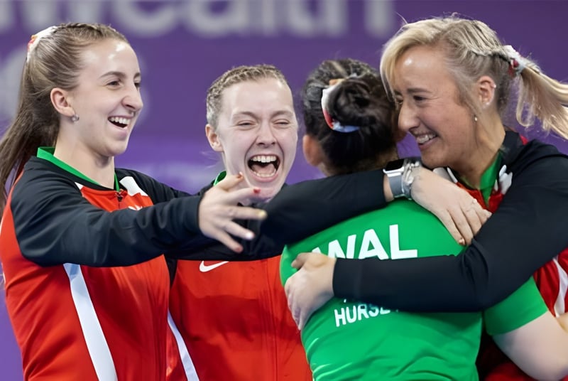 Drei Schülerinnen in Sportuniformen feiern und umarmen sich auf dem Campus der St. Michael’s School vor einem lila Hintergrund.