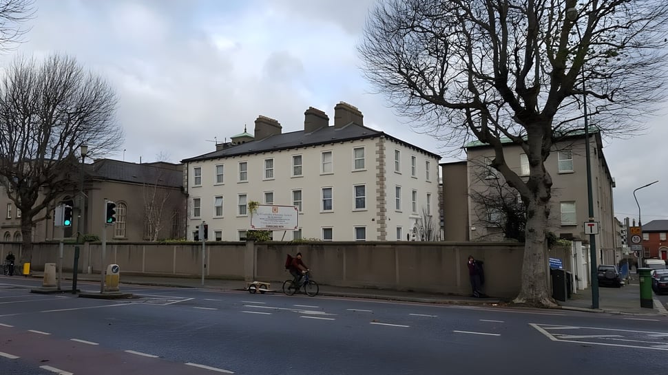 Das mehrstöckige Gebäude des St. Mogues College ist von kahlen Bäumen umgeben und eine Straße mit einem Radfahrer liegt im Vordergrund.