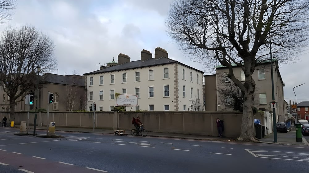 Das mehrstöckige Gebäude des St. Mogues College ist von kahlen Bäumen umgeben und eine Straße mit einem Radfahrer liegt im Vordergrund.
