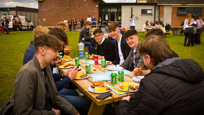 Schüler des St. Mogues College essen gemeinsam draußen an einem Tisch vor einem Van und Gebäuden.