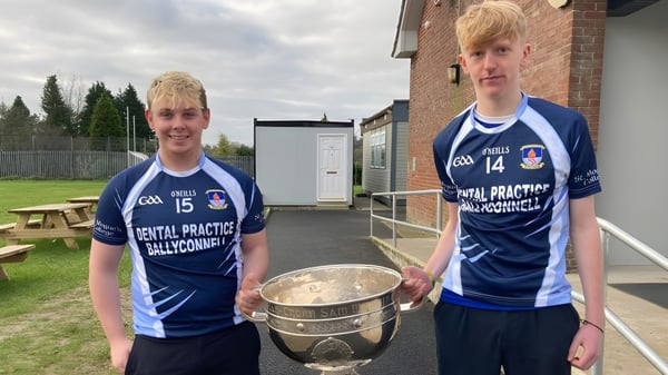 Zwei Schüler des St. Mogues College halten einen Pokal vor einem Backsteingebäude und Bäumen.