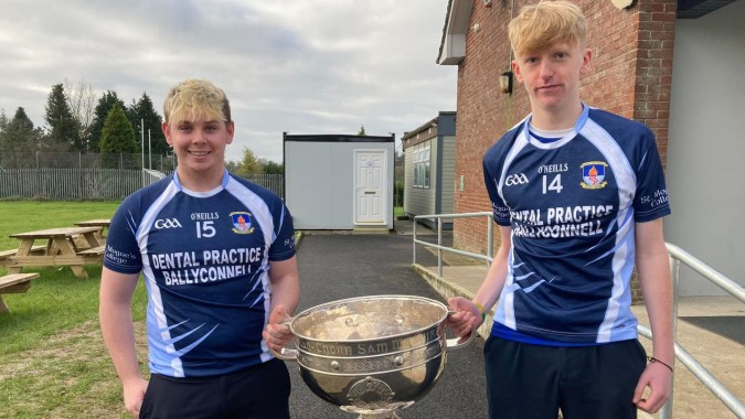 Zwei Schüler in Sportuniformen halten eine Trophäe vor dem Gebäude des St. Mogues College.