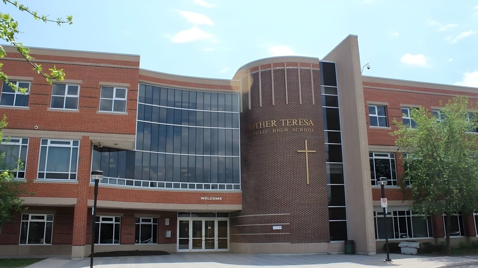 Das mehrstöckige Gebäude der St. Mother Teresa High School mit einer geschwungenen Glasfassade ist von Bäumen umgeben.