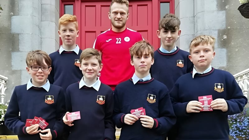 Eine Gruppe Schüler in Schuluniformen steht vor einem Gebäude mit roter Tür auf dem Campus von St. Muredach's College, begleitet von einem Mann in rotem Trikot.