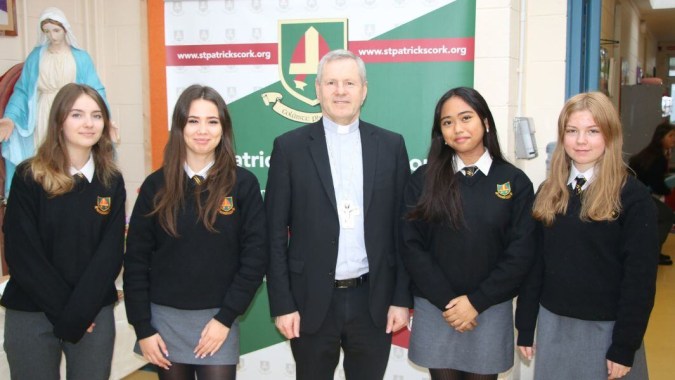 Eine Gruppe Schülerinnen und Schüler in Hogwarts-ähnlichen Schuluniformen steht mit einem älteren Mann vor einem Logo auf dem Campus von St. Patricks College (Cork).