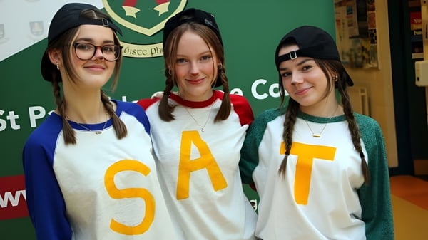 Drei Schülerinnen des St. Patricks College in weißen SAT-Shirts stehen vor grünem Hintergrund mit Wappen.