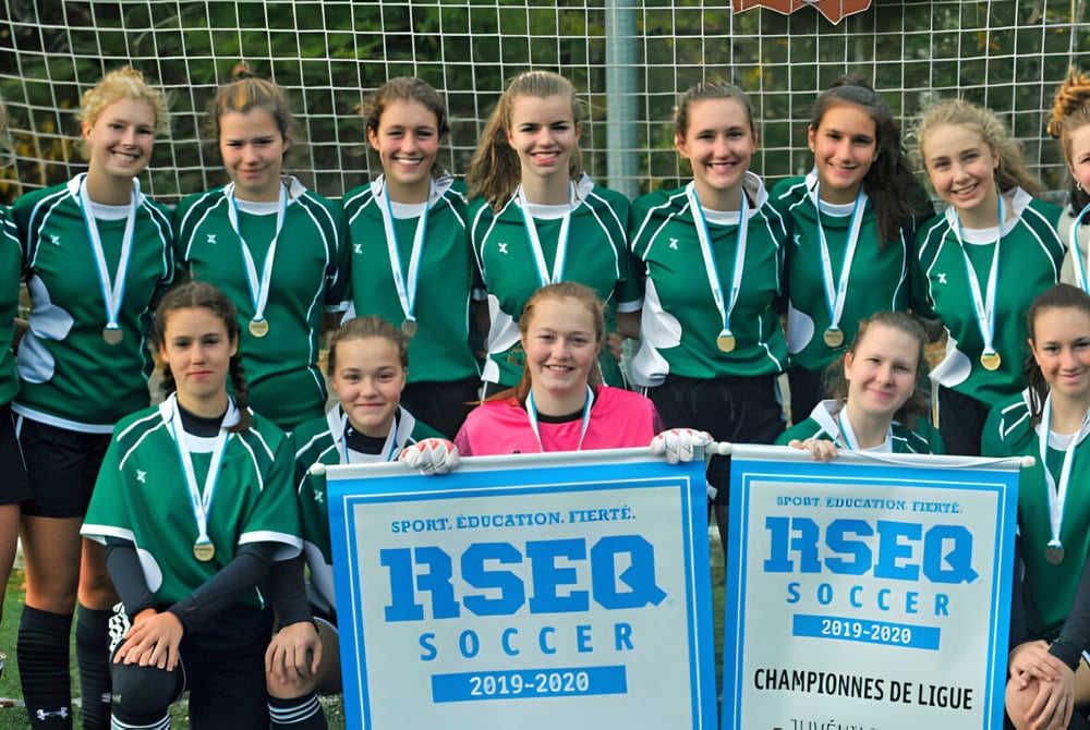 Schülerinnen der St. Patrick’s High School posieren mit Medaillen vor einem Banner der RSEQ Soccer Champions de Ligue.
