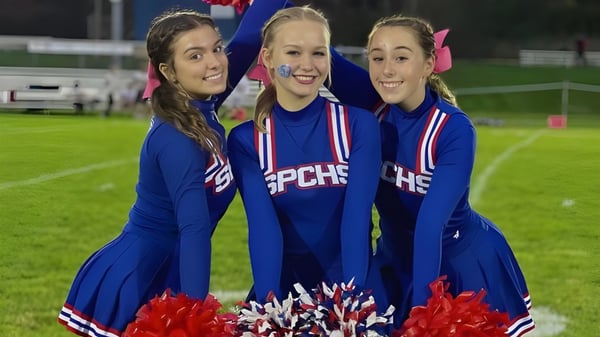 Drei Schülerinnen der St. Paul Catholic High School posieren in Cheerleading-Uniformen mit Pompons auf dem Sportplatz bei Nacht.
