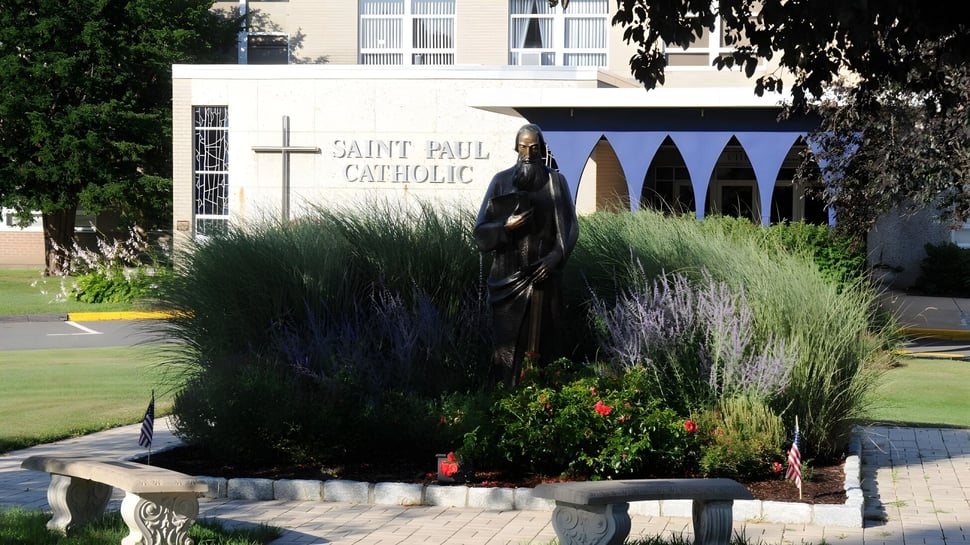 Eine Statue einer religiösen Figur steht im gepflegten Garten auf dem Gelände der St. Paul Catholic High School mit der katholischen Kirche im Hintergrund.