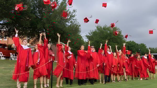 Absolventen der St. Paul High School in roten Roben stehen mit erhobenen Armen während der Abschlussfeier vor Bäumen.