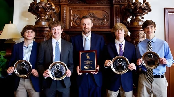 Eine Gruppe Männer in Anzügen hält Auszeichnungen vor einer verzierten Holzwand auf dem Gelände der St. Paul High School.
