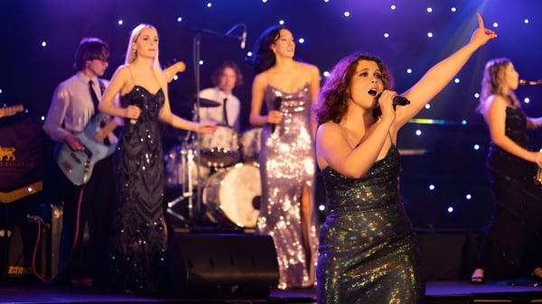 Eine Gruppe weiblicher Darstellerinnen der St Paul’s School singt und tanzt auf der Bühne mit funkelnden Lichtern im Hintergrund.