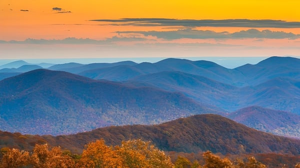 Eine herbstliche Landschaft mit bunten Bäumen und im Hintergrund sind blaue Berge bei Sonnenuntergang zu sehen.