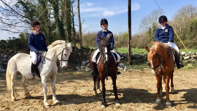 Drei Schülerinnen und Schüler der St. Paul’s Secondary in Galway reiten auf Pferden in einer ländlichen Umgebung.