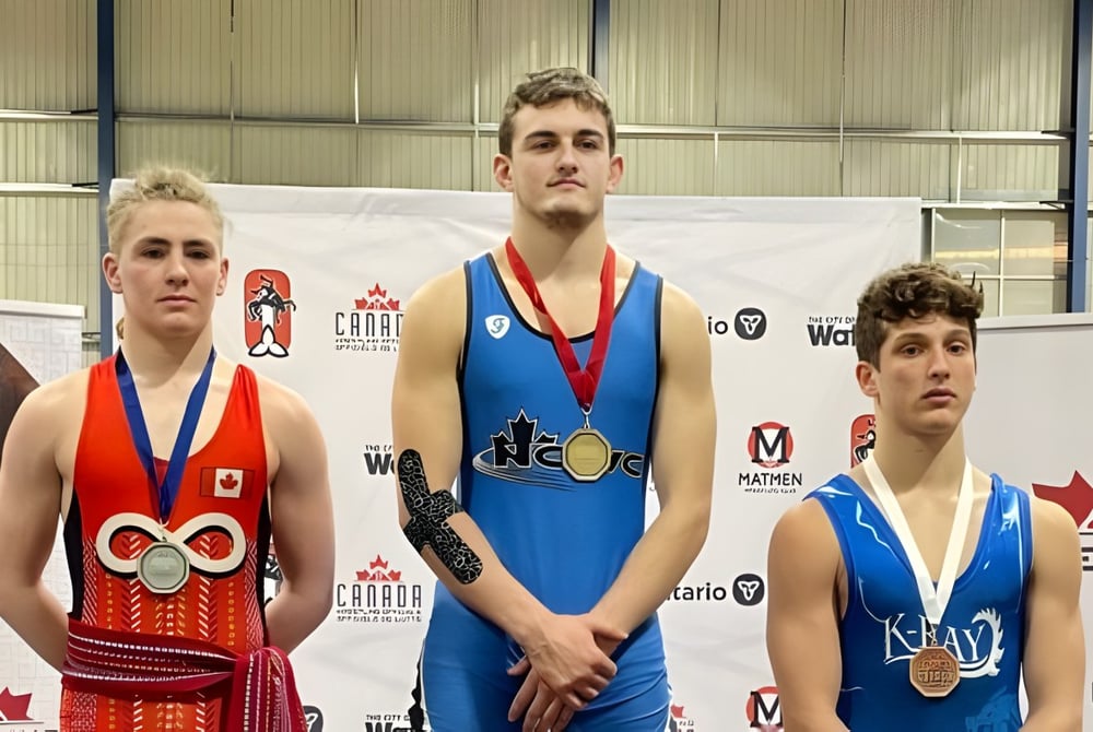 Drei Sportler mit Medaillen stehen vor einem Banner auf dem Gelände der St. Peter Catholic High School.