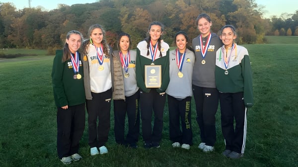 Eine Gruppe junger Athletinnen der St. Peter High School steht mit Medaillen auf einem Rasenfeld vor Bäumen.