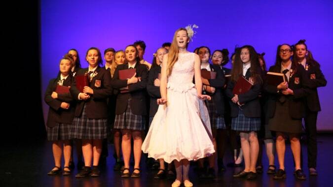 Eine Frau im weißen Kleid steht auf der Bühne vor einer Gruppe Schülerinnen in Schuluniformen auf dem Campus der St. Peter's School Cambridge.
