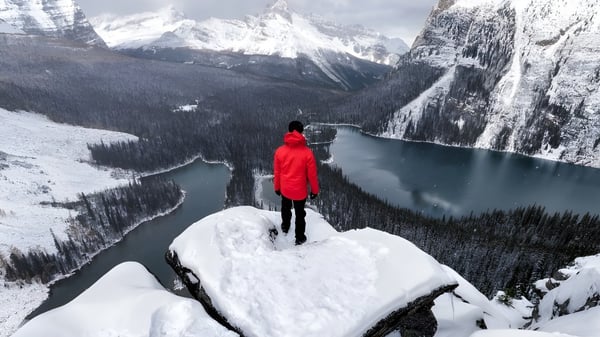 Eine Person in roter Jacke steht auf einer verschneiten Klippe mit Blick auf einen gefrorenen See und schneebedeckte Berge bei der St. Pius X High School.