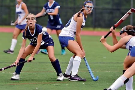Schülerinnen der St. Raphaela’s Secondary School spielen ein Hockeyspiel auf einem Spielfeld mit grünem Gras und Bäumen im Hintergrund.