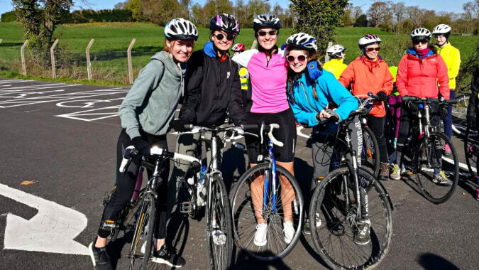 Eine Gruppe von Schülerinnen und Schülern der St. Raphaela’s Secondary School posiert in bunter Fahrradkleidung auf einer Straße neben einem Feld.