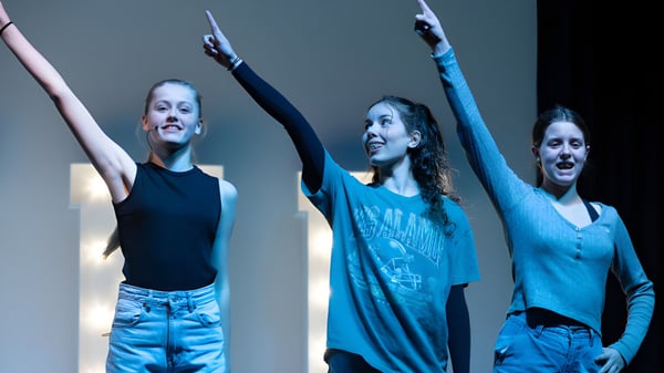 Drei Schülerinnen mit ausdrucksstarken Gesichtsausdrücken und erhobenen Armen am St. Teresa’s College vor dunklem Hintergrund.