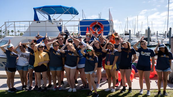 Schülerinnen der St. Thomas Aquinas High School posieren in einheitlichen Sportuniformen vor einer Marina mit Booten.