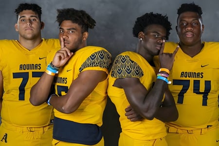 Vier Schüler der St. Thomas Aquinas High School tragen gelbe Aguias-Trikots und stehen zusammen vor dunklem Hintergrund.