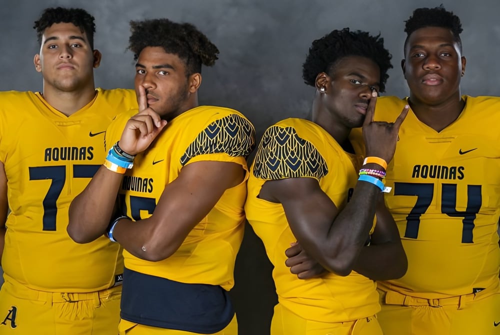 Vier Schüler der St. Thomas Aquinas High School tragen gelbe Aguias-Trikots und stehen zusammen vor dunklem Hintergrund.
