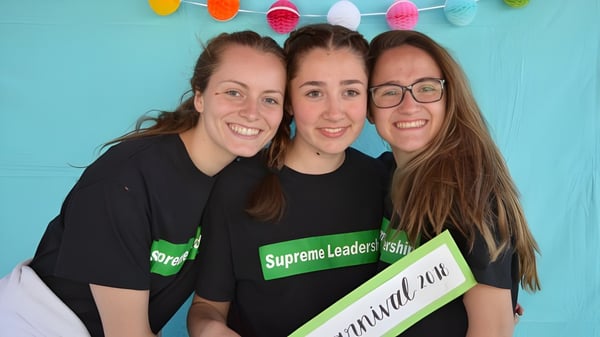 Drei Schülerinnen der St. Thomas High School lächeln und posieren vor einer türkisfarbenen Wand mit bunten Girlanden.