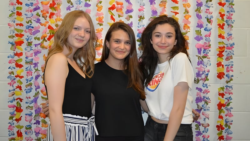 Drei Schülerinnen der St. Thomas High School stehen vor einem farbenfrohen Blumen-Vorhang.