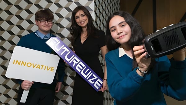 Drei Schülerinnen der St. Tiernan’s Community School stehen vor einer gemusterten Wand mit einem Schild 'Innovative'.