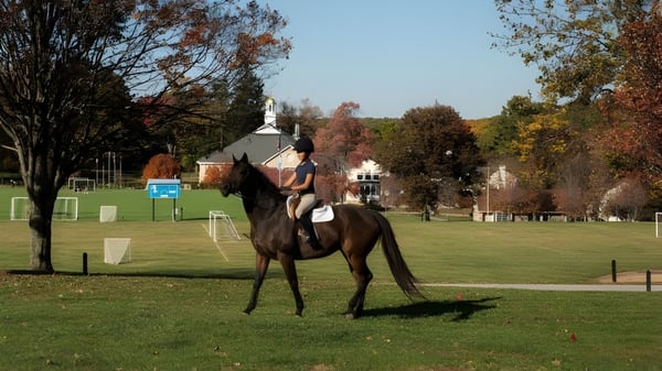 Eine Schülerin der St. Timothy's School reitet auf einem Pony auf einem grasbewachsenen Feld vor herbstlichen Bäumen.