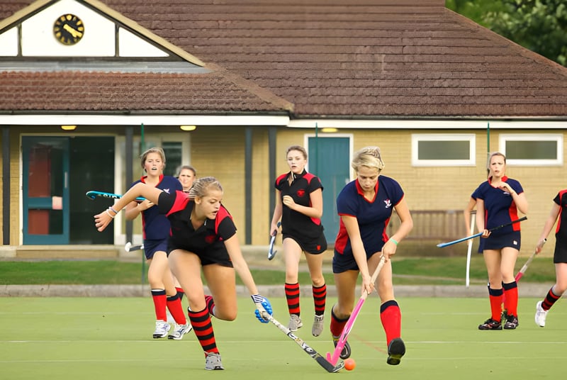 Eine Gruppe weiblicher Schülerinnen der Stamford Endowed Schools spielt Feldhockey auf einem Rasenplatz vor einem traditionellen Gebäude.