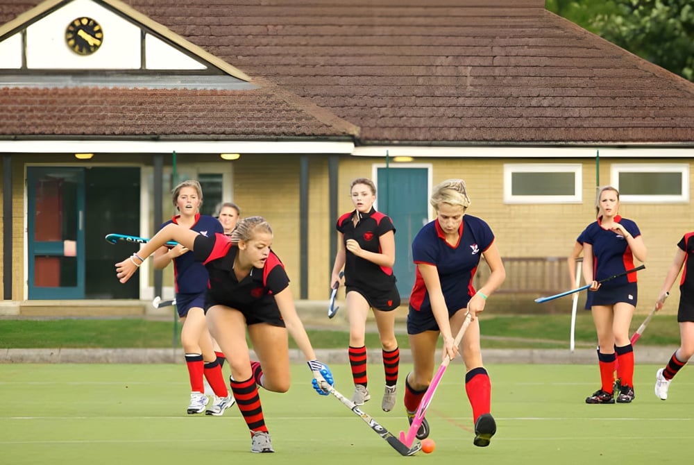 Eine Gruppe weiblicher Schülerinnen der Stamford Endowed Schools spielt Feldhockey auf einem Rasenplatz vor einem traditionellen Gebäude.