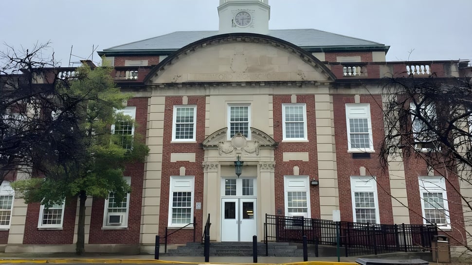 Das Backsteingebäude mit Uhrenturm der Stamford High School ist von kahlen Bäumen und einem schmiedeeisernen Zaun umgeben.