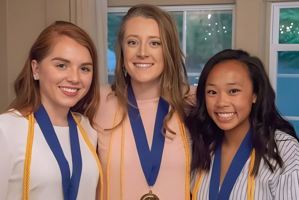 Drei Absolventinnen der Stearns High School in Abschlusskleidung halten Medaillen vor einem Fenster mit Grünblick.
