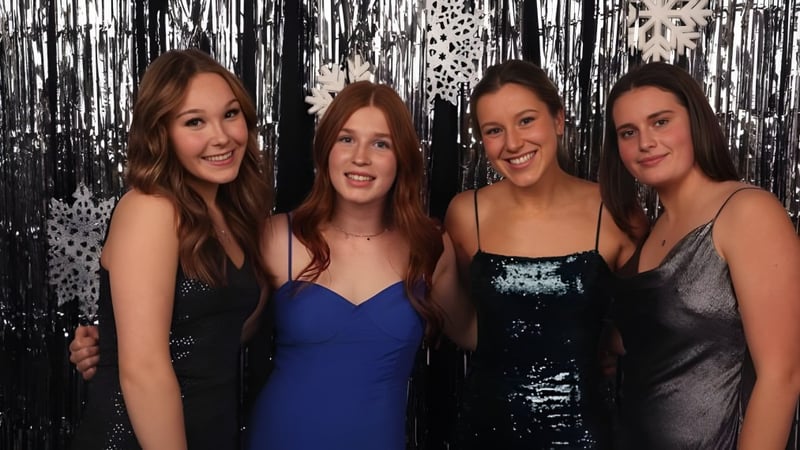 Vier Schülerinnen mit langen dunklen Haaren posieren vor einem metallischen Hintergrund auf dem Campus der Stellys Secondary School.