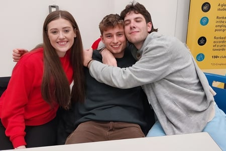 Drei Schülerinnen und Schüler umarmen sich und lächeln auf dem Campus der Stepaside Educate Together Secondary School vor Informationswänden.