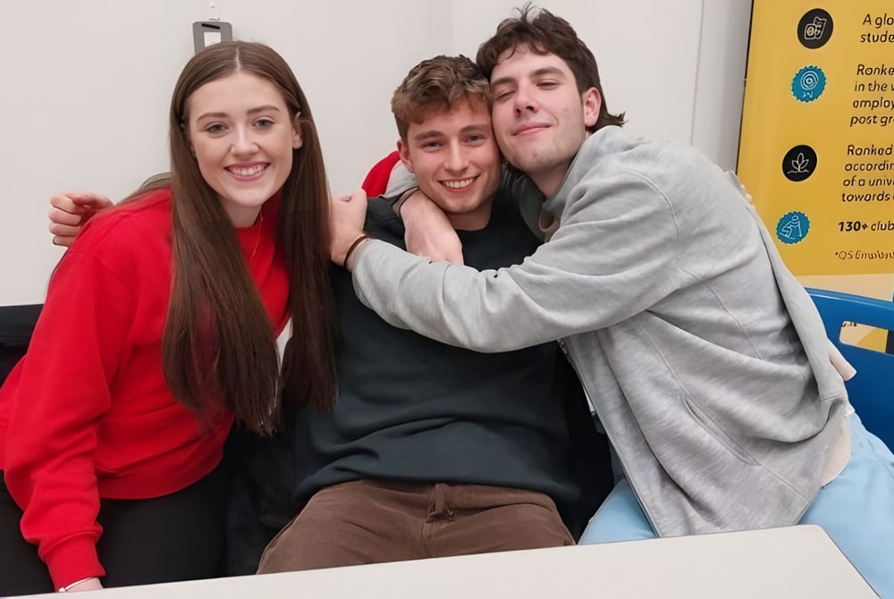 Drei Schülerinnen und Schüler umarmen sich und lächeln auf dem Campus der Stepaside Educate Together Secondary School vor Informationswänden.