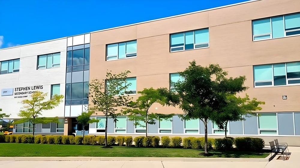 Das mehrstöckige Gebäude der Stephen Lewis Secondary School mit großen Fenstern steht vor klarem blauem Himmel und Bäumen.
