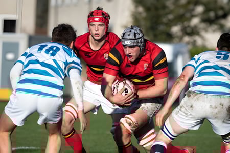 Schüler des Stewart’s Melville College spielen ein intensives Rugby-Match auf einem grasbewachsenen Spielfeld mit Gebäuden im Hintergrund.