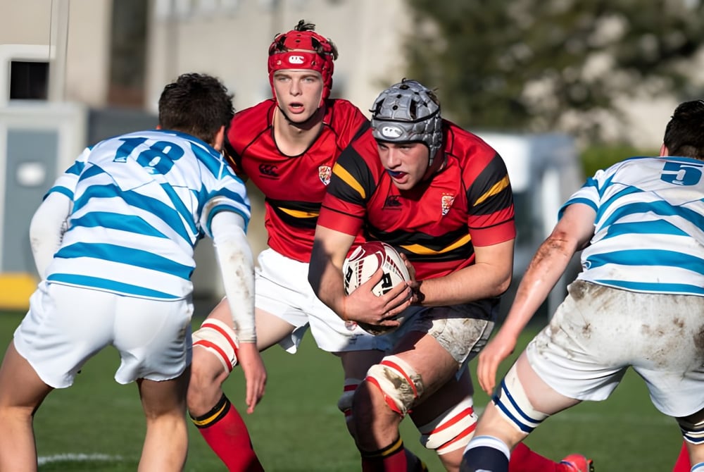 Schüler des Stewart’s Melville College spielen ein intensives Rugby-Match auf einem grasbewachsenen Spielfeld mit Gebäuden im Hintergrund.