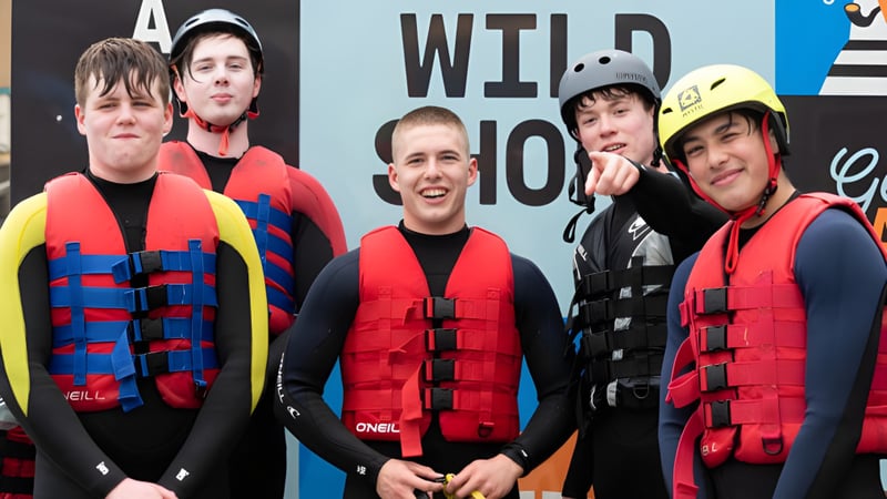 Eine Gruppe Schüler des Stewart’s Melville College trägt Schwimmwesten und Helme vor einem Schild mit der Aufschrift WILD 100.