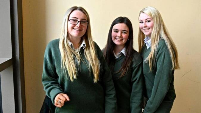 Drei Schülerinnen in grüner Schuluniform stehen im Korridor der St. Mary's Holy Faith (Killester) und lächeln zusammen.